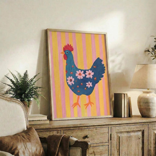 May include: Framed folk art poster featuring a blue hen with red comb and pink floral pattern on body, set against yellow and pink striped background. Artwork displayed on rustic wooden sideboard next to fern plant, books, and ceramic vases in neutral-toned living room setting.