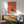 May include: Modern orange and teal geometric poster artwork displayed in wood frame above mid-century credenza. Features minimalist illustration of person reading by window with moon visible. Shown with sage green upholstered armchair, white ceramic table lamp, and round coffee table in sunlit room with white walls and natural shadows.