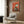 May include: Modern living room with beige armchair and patterned pillow, brass floor lamp, dark round side table with open book, wood paneled wall. Framed poster shows black and white tuxedo cat sitting on turquoise wooden chair with small dark birds perched on chair back and ground, set against textured coral orange background.