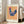 May include: Framed folk art poster featuring a stylized blue hen with red comb and wattle, decorated with pink flowers across its body, standing on orange legs against yellow and pink vertical stripes. The artwork is displayed in a light wood frame with white matting, leaning against a beige sofa in a bright living room with large windows and natural lighting.