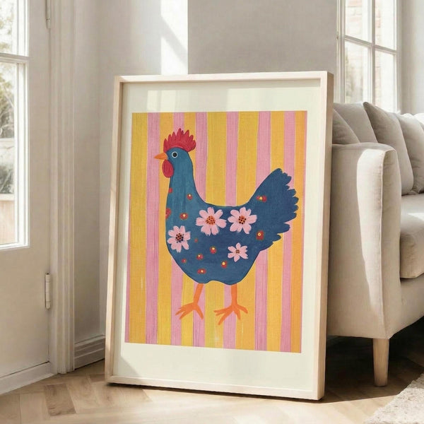 May include: Framed folk art poster featuring a stylized blue hen with red comb and wattle, decorated with pink flowers across its body, standing on orange legs against yellow and pink vertical stripes. The artwork is displayed in a light wood frame with white matting, leaning against a beige sofa in a bright living room with large windows and natural lighting.