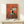 May include: Framed poster print displayed on light wood shelf showing black and white tuxedo cat sitting on turquoise wooden chair against coral red textured background, with several small dark birds perched on chair back and floor. Small white gift box with greenery sits beside gold-framed artwork on minimalist wooden furniture against cream wall.