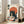 May include: Framed poster artwork in light wood frame leaning against white wall next to beige sofa in bright living room. Shows Japanese-style illustration of black cat with white eyes eating ramen noodles with chopsticks, sitting behind large bowl with fried egg. Orange sun circle backdrop, cream background with Japanese text. Vintage ukiyo-e art style with orange, black, cream colors. Small sauce bottles visible beside bowl.