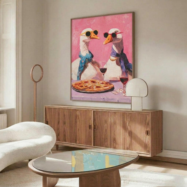 May include: Framed artwork showing two white geese wearing sunglasses and colorful scarves sitting at a table with pizza and wine glasses, displayed above a wooden credenza with brass handles. Modern living room setting features a white curved chair, oval glass coffee table, and neutral beige walls with natural lighting.
