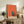 May include: Framed contemporary art print with orange and coral tones showing a small window with blue sky and silhouetted figure, displayed on rustic wooden sideboard alongside fern plant, ceramic vase, and books in warm neutral living room setting