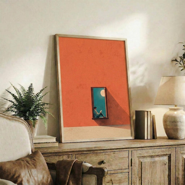May include: Framed contemporary art print with orange and coral tones showing a small window with blue sky and silhouetted figure, displayed on rustic wooden sideboard alongside fern plant, ceramic vase, and books in warm neutral living room setting