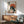 May include: Japanese-style poster print featuring a black cat eating ramen noodles from a decorative bowl, set against a large orange sun with Japanese text characters. The artwork shows vintage ukiyo-e styling in cream, orange, and black colors, displayed in a wooden frame above a mid-century modern walnut credenza. Scene includes a teal upholstered armchair, white ceramic lamp, and warm natural lighting on white walls.