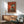 May include: Modern living room with wooden sideboard, teal upholstered chair, and white table lamp. Framed artwork on white wall shows black and white tuxedo cat sitting on turquoise wooden chair with several small dark birds perched above and below, set against warm reddish-orange textured background. Natural sunlight creates geometric shadows across the scene.