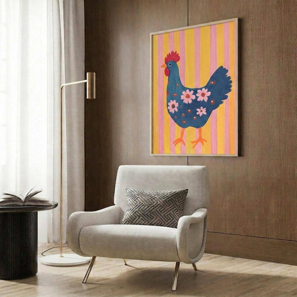 May include: Framed poster print featuring a decorative blue hen with red comb and pink floral details against yellow and pink vertical stripes, displayed on wood-paneled wall above beige upholstered armchair with geometric pillow, brass floor lamp and side table nearby in modern living room setting