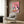 May include: Whimsical artwork featuring two white geese wearing sunglasses seated at a table with pizza and red wine against a pink background, displayed in light wood frame on dark wood wall paneling. Modern beige armchair with geometric patterned pillow, brass floor lamp, and dark side table with open book complete the contemporary living room setting.