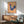 May include: Framed folk art poster featuring a blue hen with red comb and pink floral patterns on body, displayed against yellow and pink striped background. Shown hanging above mid-century modern walnut credenza with white ceramic lamp, next to sage green upholstered armchair in bright, naturally-lit room with white walls.