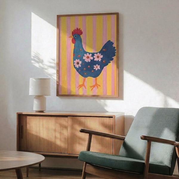 May include: Framed folk art poster featuring a blue hen with red comb and pink floral patterns on body, displayed against yellow and pink striped background. Shown hanging above mid-century modern walnut credenza with white ceramic lamp, next to sage green upholstered armchair in bright, naturally-lit room with white walls.