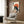 May include: Japanese-style poster print displayed on wood-paneled wall above beige upholstered armchair. Artwork features black cat eating ramen noodles with chopsticks against orange sun background, with Japanese text and traditional ukiyo-e art style. Modern living room setting includes geometric patterned pillow, brass floor lamp, and dark side table with open book.