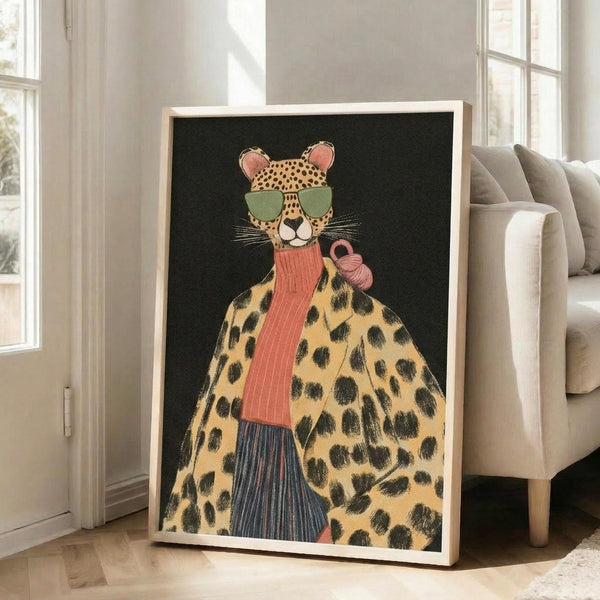 May include: Large framed poster leaning against white wall next to beige sofa, showing stylized leopard wearing green sunglasses, coral ribbed scarf, and leopard print coat against dark background. Room has white walls, wooden floors, and natural light from windows.