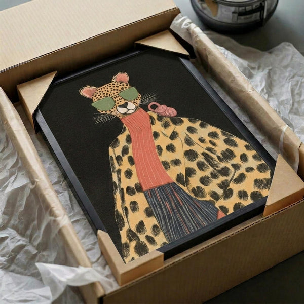 Stylized leopard wearing green sunglasses and coral turtleneck sweater illustrated on dark background, displayed in black frame being unboxed from cardboard shipping package with protective wrapping materials and tape dispenser visible nearby