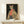 May include: Framed poster print featuring an anthropomorphic cheetah wearing green sunglasses, coral ribbed scarf, and leopard print coat against black background, displayed on light wood console table next to small white wrapped gift with evergreen sprig, against cream colored wall