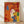 May include: Colorful stylized illustration of four dogs in a thin wooden frame, leaning against a white wall on light wood flooring. The artwork features dogs in vibrant orange, white, yellow, and blue with contrasting colored ears against warm red and yellow geometric background shapes. The print has a textured, painterly quality with visible brush-like effects throughout the composition.