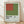 May include: Framed museum exhibition poster featuring geometric design with rust red rectangle positioned over olive green square on light blue background. Black text displays artist name and Detroit Institute of Arts with exhibition dates. Natural wood frame shown against white wall on light wooden floor with soft window shadows.
