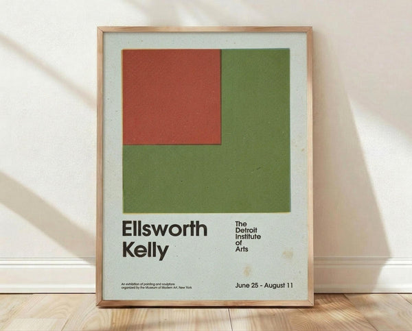 May include: Framed museum exhibition poster featuring geometric design with rust red rectangle positioned over olive green square on light blue background. Black text displays artist name and Detroit Institute of Arts with exhibition dates. Natural wood frame shown against white wall on light wooden floor with soft window shadows.