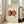 May include: Framed poster with cream background featuring overlapping geometric shapes in coral pink and black, with three small black dots below and "MODERN REAL ESTATE" text at top. Light wood frame leaning against white wall next to beige upholstered chair in bright, airy room with large windows and natural lighting.