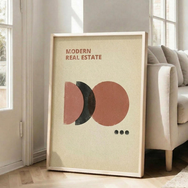 May include: Framed poster with cream background featuring overlapping geometric shapes in coral pink and black, with three small black dots below and "MODERN REAL ESTATE" text at top. Light wood frame leaning against white wall next to beige upholstered chair in bright, airy room with large windows and natural lighting.