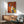 May include: Colorful abstract dog illustration artwork in wooden frame hanging above mid-century modern walnut credenza with white ceramic lamp, next to sage green upholstered armchair. Artwork features four stylized dog heads in warm orange, red, yellow, white, and blue tones against textured background. Sunlight streams across white wall creating geometric shadows in modern living room setting.