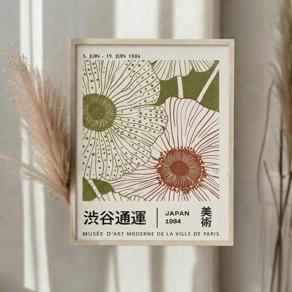 May include: Framed vintage-style Japanese exhibition poster featuring two large stylized flowers - one green daisy-like bloom and one coral-colored sunflower with detailed line work and dots. Japanese text and "JAPAN 1984" visible at bottom. Light wood frame displayed on neutral wall with dried pampas grass stems creating soft shadows in natural lighting.