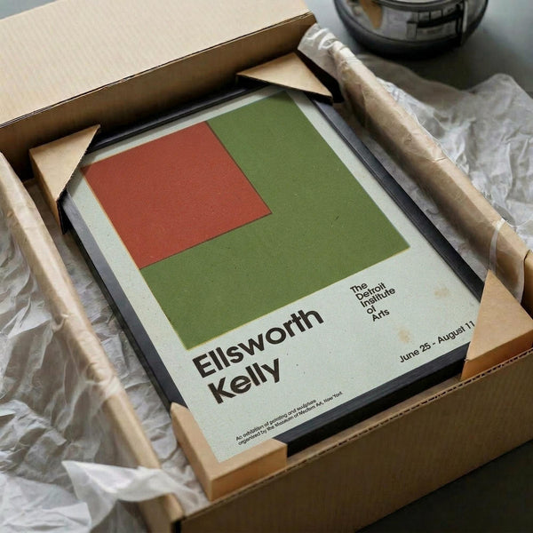 May include: Framed art print featuring abstract geometric composition with olive green rectangle and smaller coral red square on cream background. Black text shows artist name and exhibition details. Print displayed in black frame inside cardboard shipping box with protective wrapping materials and wooden support pieces.