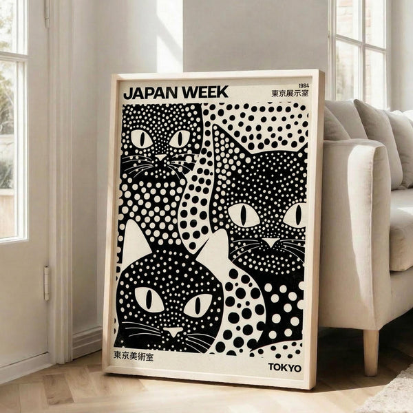 May include: A framed black and white poster featuring stylized cat faces with polka dot patterns in varying sizes. The artwork displays bold geometric designs with white dots on black backgrounds, creating striking cat silhouettes with prominent eyes and whiskers. Text reads "JAPAN WEEK" at top and "TOKYO" at bottom with Japanese characters. The light wood frame leans against a white wall next to a beige upholstered chair in a bright, minimally decorated room with hardwood floors and la