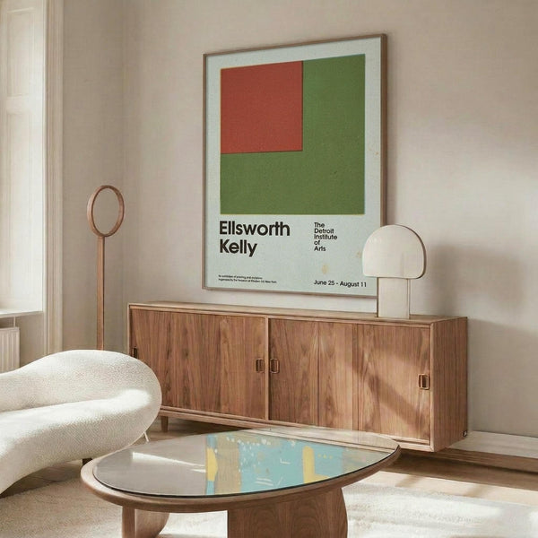 May include: Minimalist exhibition poster with green and red geometric shapes displayed in thin wooden frame above walnut credenza. Modern living room setting with cream upholstered chair, oval glass coffee table, and neutral walls. Poster features bold color blocking and clean typography against white background.