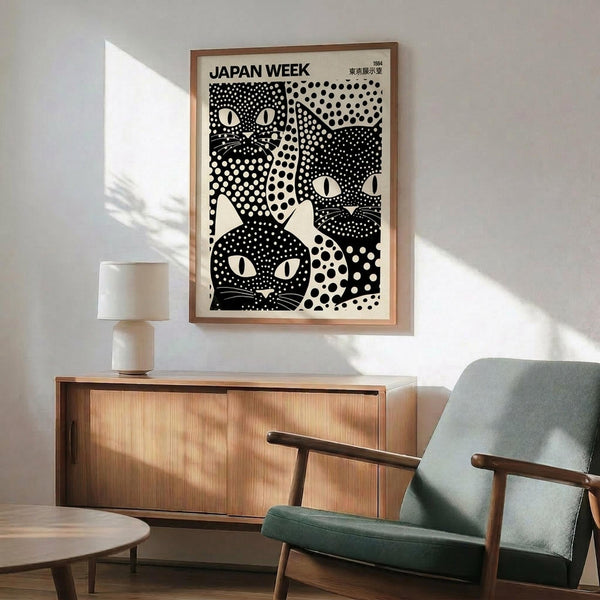 May include: Black and white poster featuring stylized cats with polka dot patterns in a wooden frame, hanging above a mid-century modern wooden credenza with white ceramic lamp, next to a sage green upholstered armchair with wooden arms in a sunlit room with white walls.