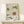 May include: Framed vintage-style Japanese art exhibition poster featuring stylized flowers in green and coral colors with decorative patterns, displayed on light wood furniture next to a small wrapped gift with evergreen sprig against a cream wall.
