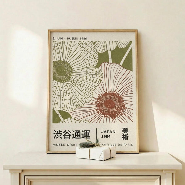 May include: Framed vintage-style Japanese art exhibition poster featuring stylized flowers in green and coral colors with decorative patterns, displayed on light wood furniture next to a small wrapped gift with evergreen sprig against a cream wall.