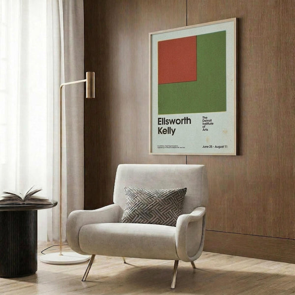 May include: Framed Ellsworth Kelly exhibition poster featuring red square on green background, displayed on wood-paneled wall above modern cream upholstered chair with geometric pillow. Brass floor lamp and side table with open book visible. Minimalist contemporary room setting with neutral tones and natural lighting from window with curtains.
