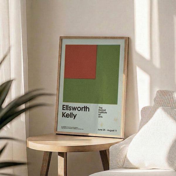 May include: Framed exhibition poster featuring green and red geometric color blocks with black text reading "Ellsworth Kelly" and "The Detroit Institute of Arts" with exhibition dates June 25 - August 11. Displayed on light wood round side table next to beige upholstered chair with plant leaves visible, natural sunlight streaming across neutral wall creating soft shadows.