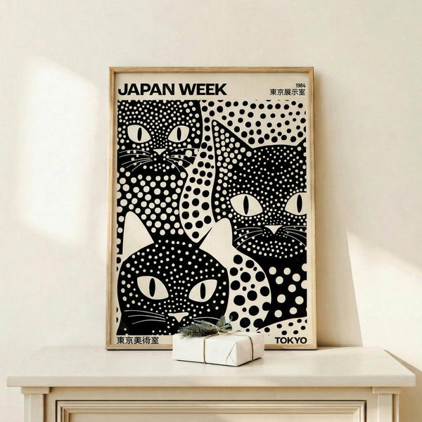 May include: Black and white poster in light wood frame showing stylized cats with polka dot patterns and large eyes, displayed on pale wooden shelf next to small wrapped gift with dried botanical sprig, against cream colored wall