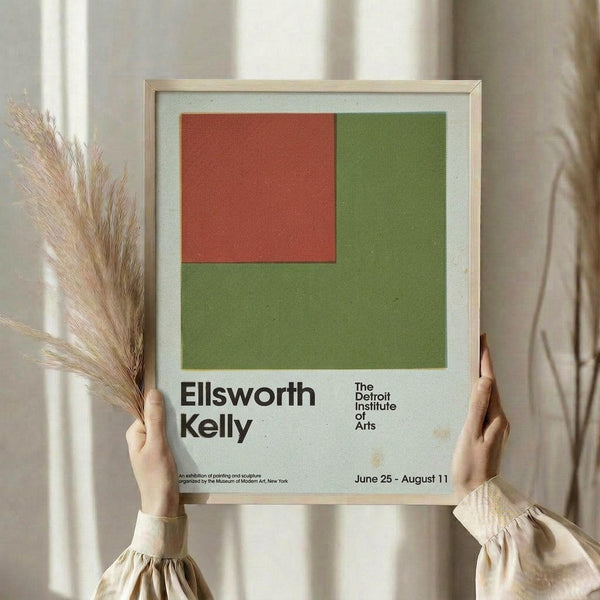 May include: Person holding framed vintage exhibition poster featuring geometric color blocks in orange-red and olive green with black text reading "Ellsworth Kelly" and "The Detroit Institute of Arts" with exhibition dates. Light wood frame against white curtained window with dried pampas grass visible. Minimalist home decor styling in neutral tones.