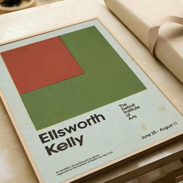 May include: Vintage exhibition poster for Ellsworth Kelly at The Detroit Institute of Arts, featuring abstract geometric blocks in rust red and olive green on pale background. Poster is matted in cream frame, positioned on white surface next to rolled papers and ceramic objects in soft natural lighting.