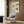 May include: Framed vintage Japanese exhibition poster with green and coral flower designs displayed on wood-paneled wall above cream upholstered chair with geometric pillow. Modern brass floor lamp and dark round side table with open book complete the warm, contemporary living space with natural lighting from window.