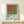 May include: Framed vintage museum exhibition poster featuring minimalist geometric design with red square and olive green shapes on light blue background. Displayed on light wood console with small wrapped gift beside it. Natural lighting creates soft shadows on cream wall.