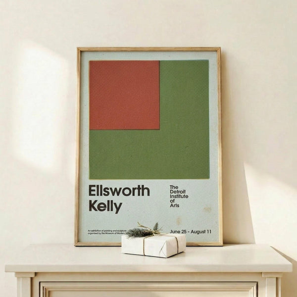 May include: Framed vintage museum exhibition poster featuring minimalist geometric design with red square and olive green shapes on light blue background. Displayed on light wood console with small wrapped gift beside it. Natural lighting creates soft shadows on cream wall.
