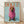 May include: Framed poster print leaning against white wall on light wood floor. Features dachshund dog wearing oversized pink knit hoodie on pale blue background. Orange "DOGUE" text at top mimics fashion magazine style. Natural wood frame surrounds the artwork. Soft shadows cast on textured white wall behind.