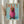 May include: Framed poster print displayed on white wall showing dachshund dog wearing oversized pink knit hoodie against light blue background, with "DOGUE" text in orange letters at top mimicking fashion magazine cover style. Dried pampas grass visible in foreground, white curtains in background.