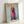 May include: Framed poster print leaning against white wall showing dachshund dog wearing oversized pink knitted hoodie on light blue background, with orange "DOGUE" text at top in magazine-style layout, displayed in natural wood frame