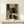 May include: Abstract geometric artwork in wooden frame leaning against white wall on light wood surface, featuring overlapping rectangular and rounded shapes in black, sage green, coral, navy blue, beige and gray with textured brushstroke effects. Small white wrapped gift box with natural twine and greenery sprig sits in front of the framed piece.