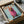 May include: Framed poster print in cardboard shipping box with protective packaging. Print shows dachshund dog wearing oversized pink knit hoodie on light blue background with orange "DOGUE" text in fashion magazine style. Black frame visible with wooden corner protectors and white tissue paper packaging materials surrounding the box.