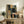 May include: Abstract geometric artwork with overlapping rectangular and rounded shapes in dark green, coral, blue, and cream tones, displayed in light wood frame on rustic wooden sideboard alongside potted fern, ceramic vase, and stacked books in cozy living room setting with neutral armchair.