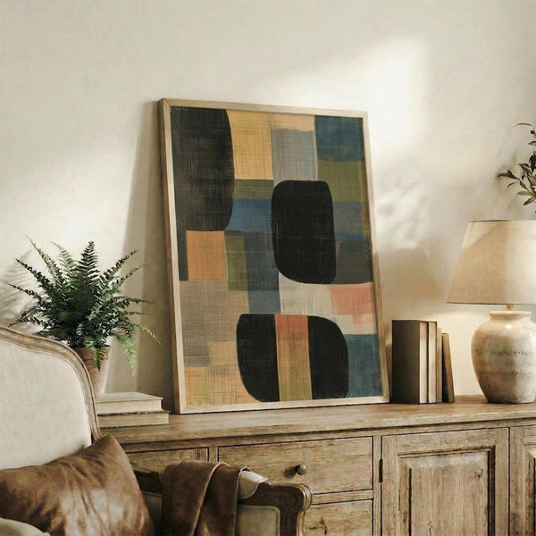 May include: Abstract geometric artwork with overlapping rectangular and rounded shapes in dark green, coral, blue, and cream tones, displayed in light wood frame on rustic wooden sideboard alongside potted fern, ceramic vase, and stacked books in cozy living room setting with neutral armchair.