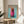 May include: Framed poster featuring a dachshund wearing a bright pink knit hoodie against a turquoise background, with "DOGUE" text in orange letters at the top. The wooden-framed print sits on a round wooden side table next to a beige upholstered chair, with a plant and white curtains visible in the sunlit room setting.