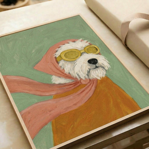 May include: A framed art print showing a white fluffy terrier dog wearing yellow aviator goggles and a flowing pink scarf against a sage green background with warm orange accents. The artwork has a painted, illustrative style and sits on white fabric with cream ribbon nearby, suggesting gift wrapping materials.